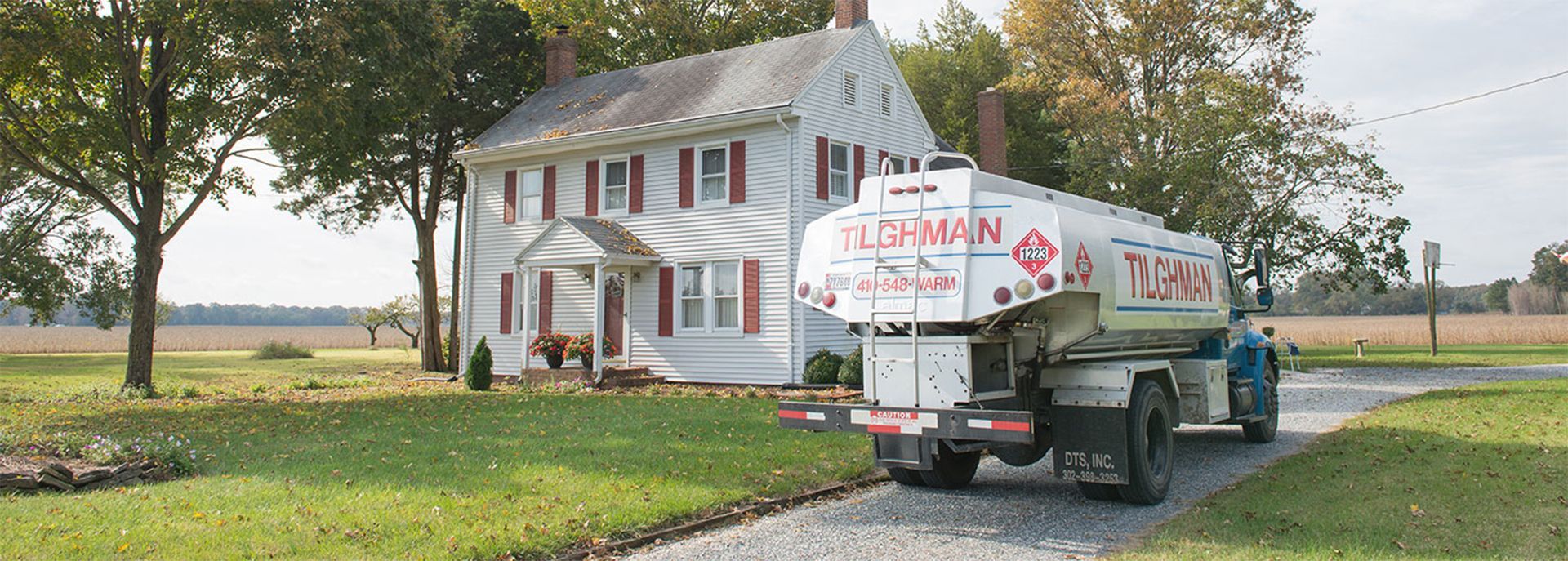 Truck carrying an old white house replica on a rural road beside a large farmhouse and trees