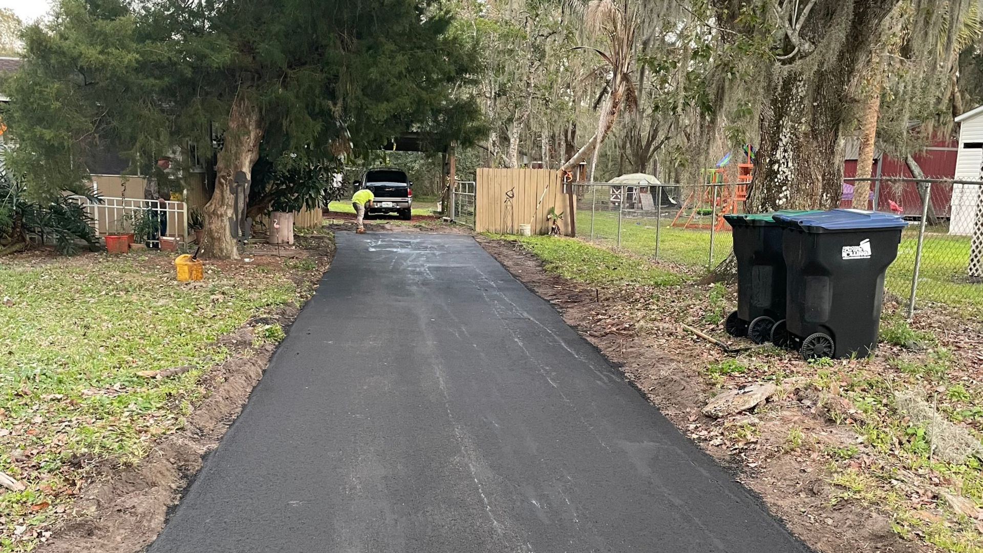 Newly paved asphalt driveway leading to a house. Green grass on either side with trees and a truck in the distance.