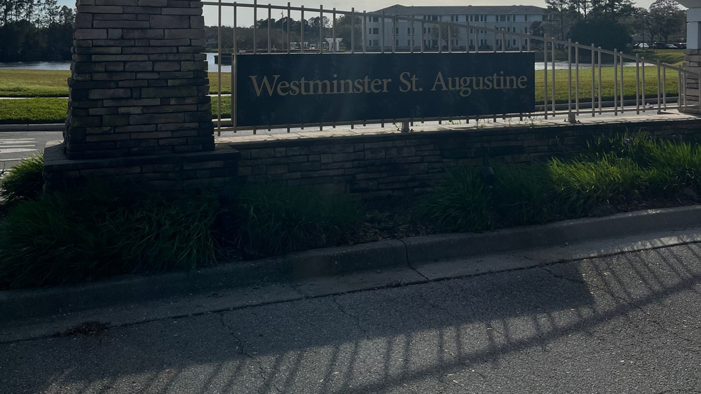 Sign for Westminster St. Augustine, a senior living community, with a brick pillar, black text, and a wrought iron fence.
