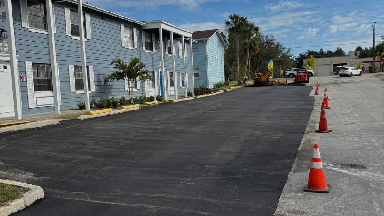 Newly paved asphalt parking lot with orange traffic cones and two-story blue apartment buildings.