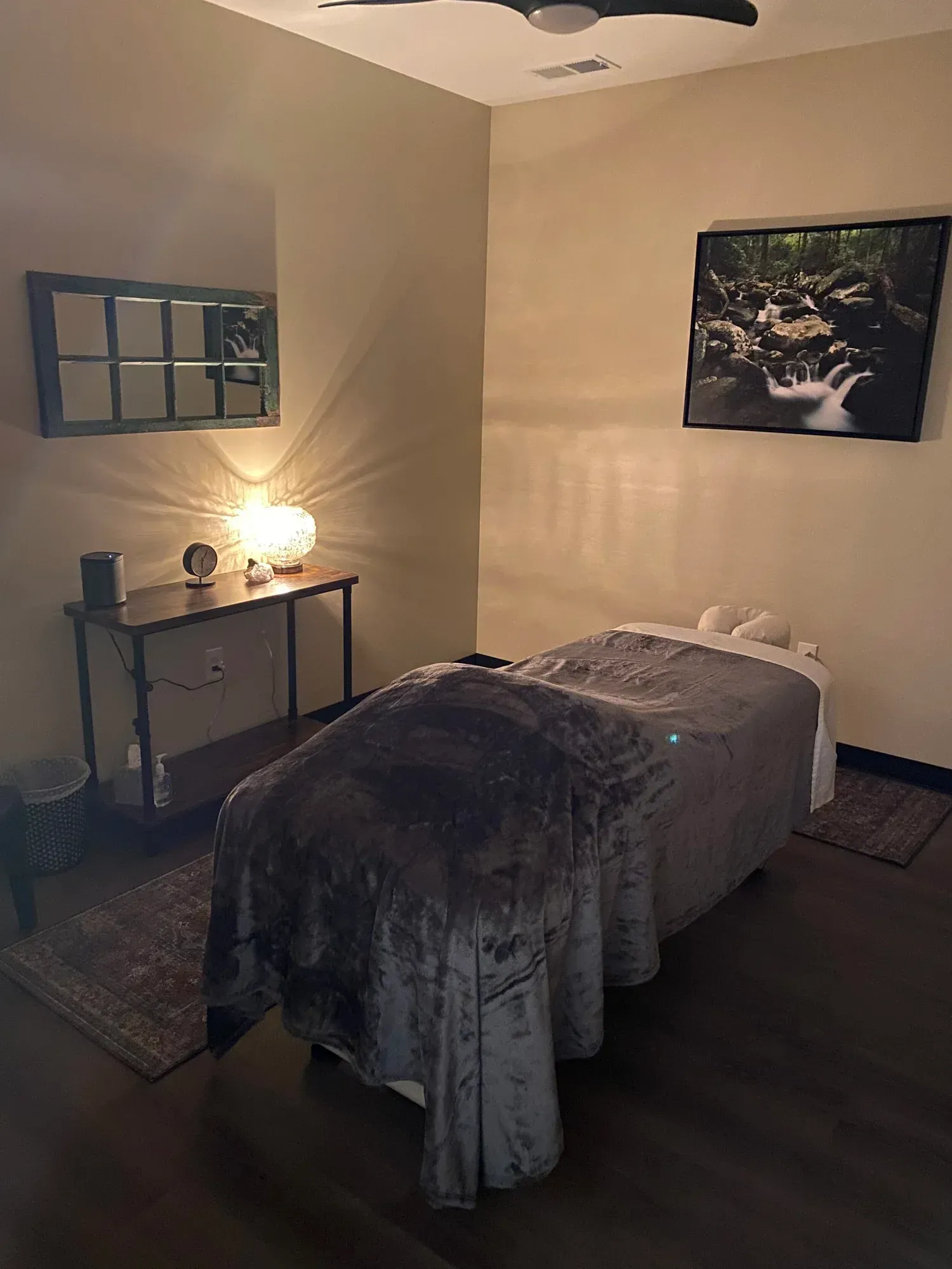 Massage room: massage table with soft blanket, table with lamp and mirror, art, soft lighting.