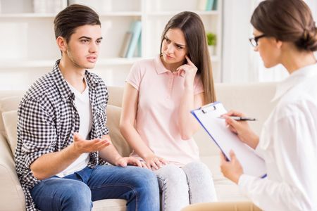 A young couple consults a therapist in a counseling session. The woman looks concerned while the man gestures.