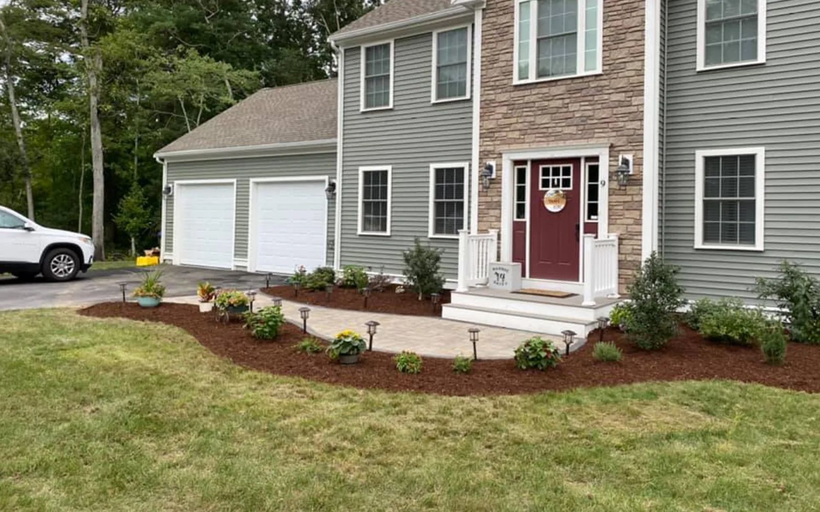 Two-story house with gray siding, a brick entryway, and a driveway lined with plants and lights.