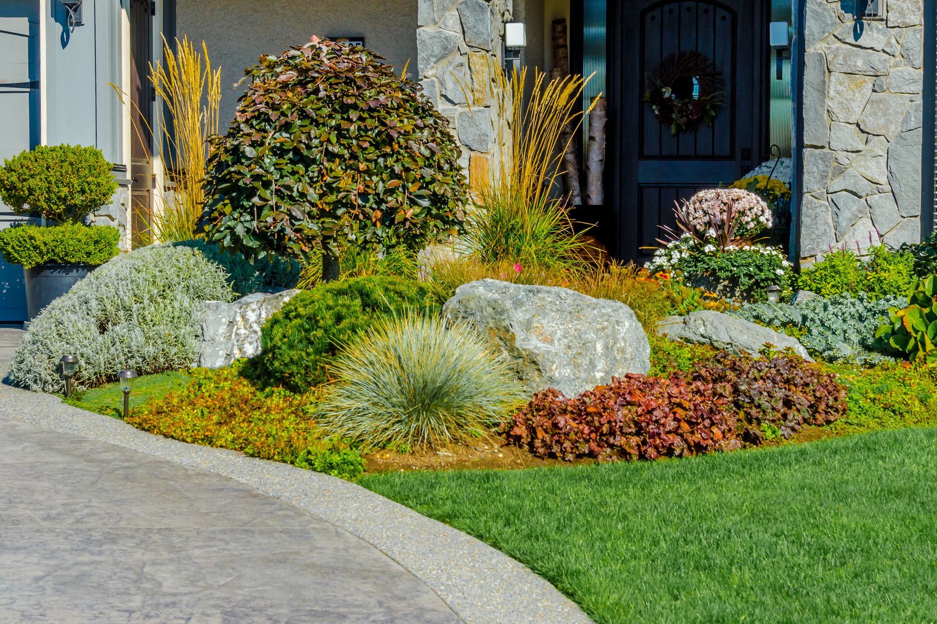 Landscaped front yard with rocks, various green and reddish plants, and a gray stone house.