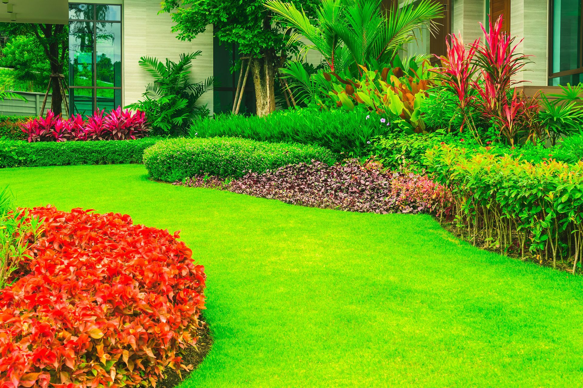 Lush green lawn with colorful landscaping, including red and pink flowering plants and hedges, in front of a house.