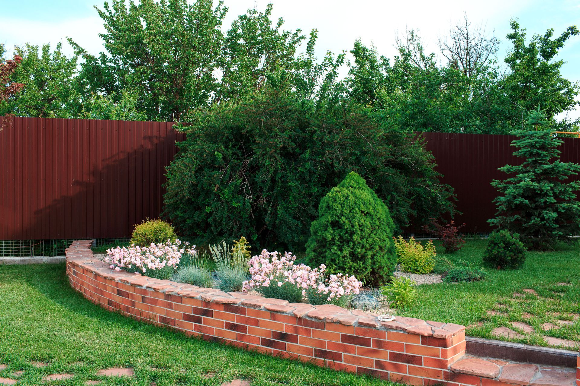 Brick-lined garden bed with flowers, shrubs, and evergreens in front of a brown fence and trees.