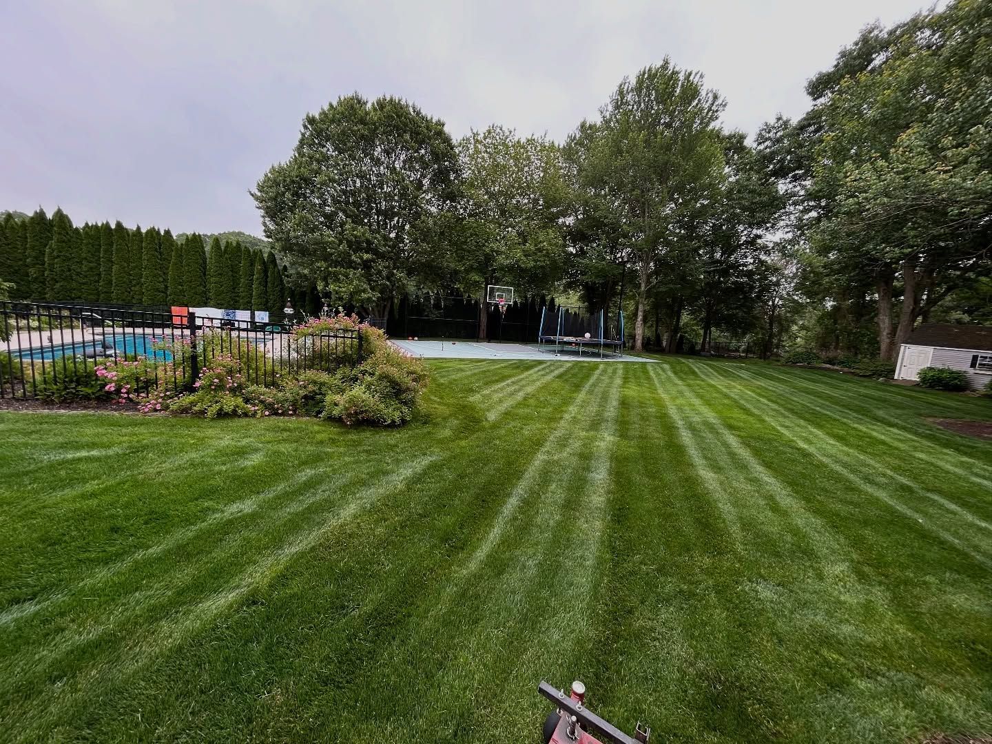 Lawn with fresh mowing stripes, trees, and a swimming pool in the background on a cloudy day.