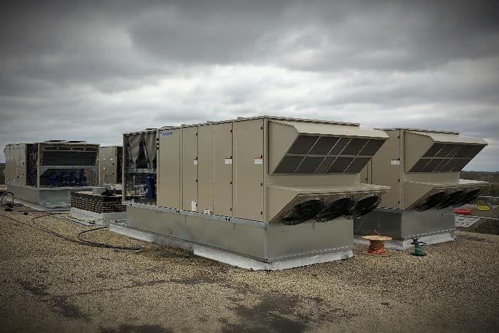 Rooftop HVAC units on a cloudy day. Gray metal boxes with vents and fans sit atop a flat, gravel-covered roof.