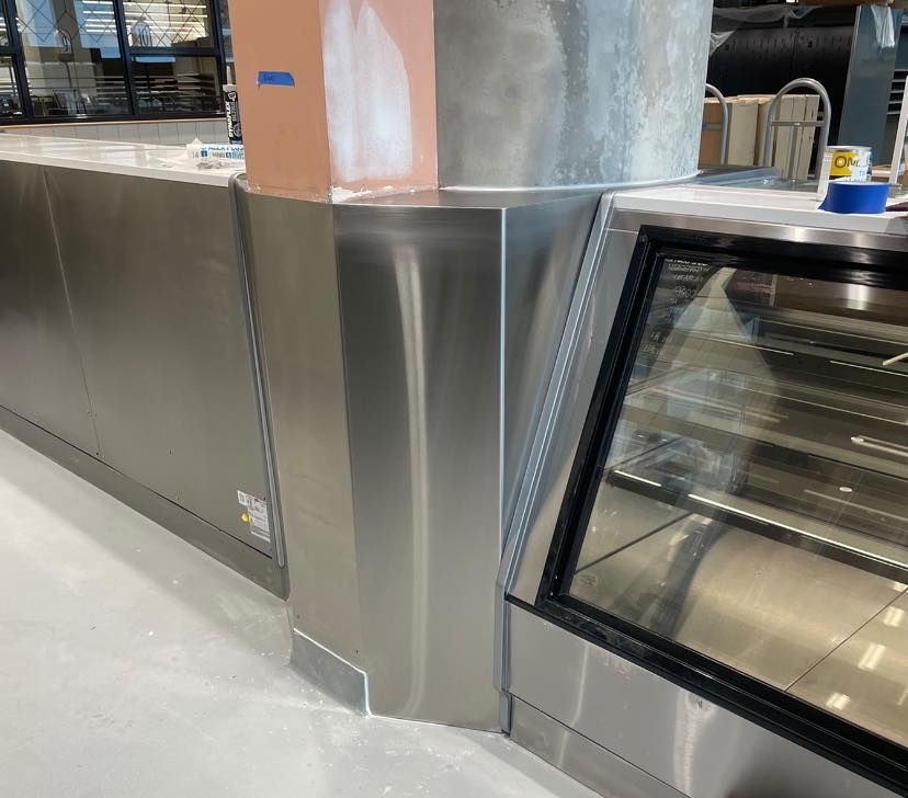 Stainless steel retail counter with display case, next to a column, in a brightly lit space.