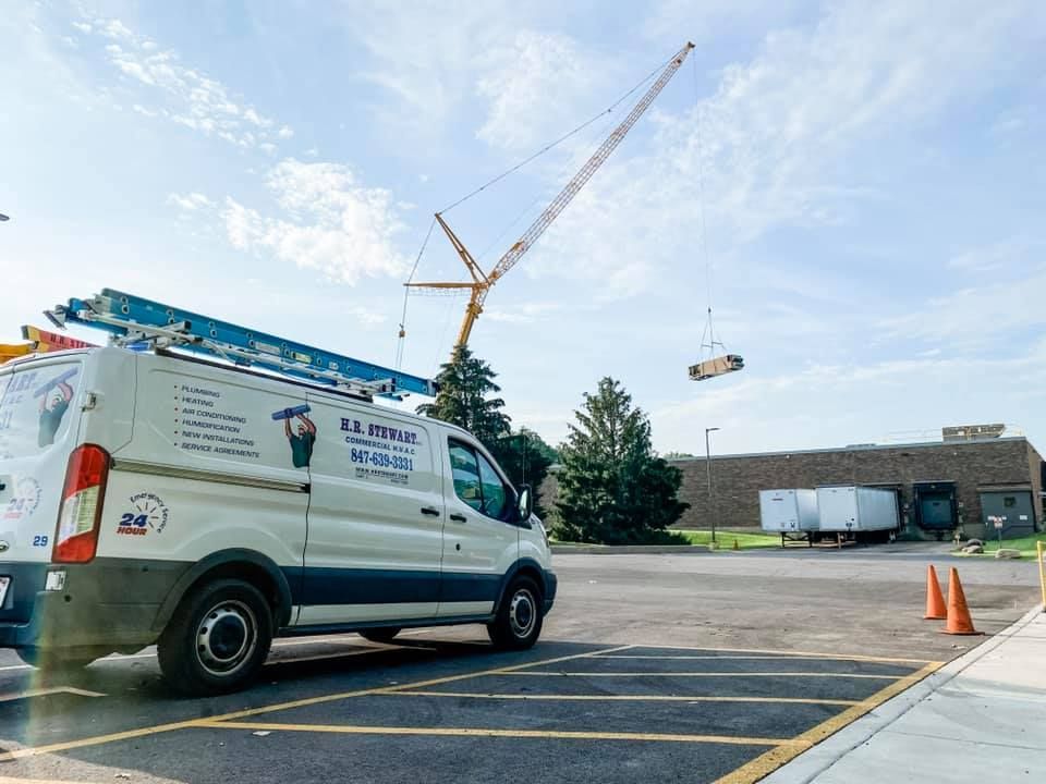 White van with company logo, crane lifting object near a building under a blue sky.