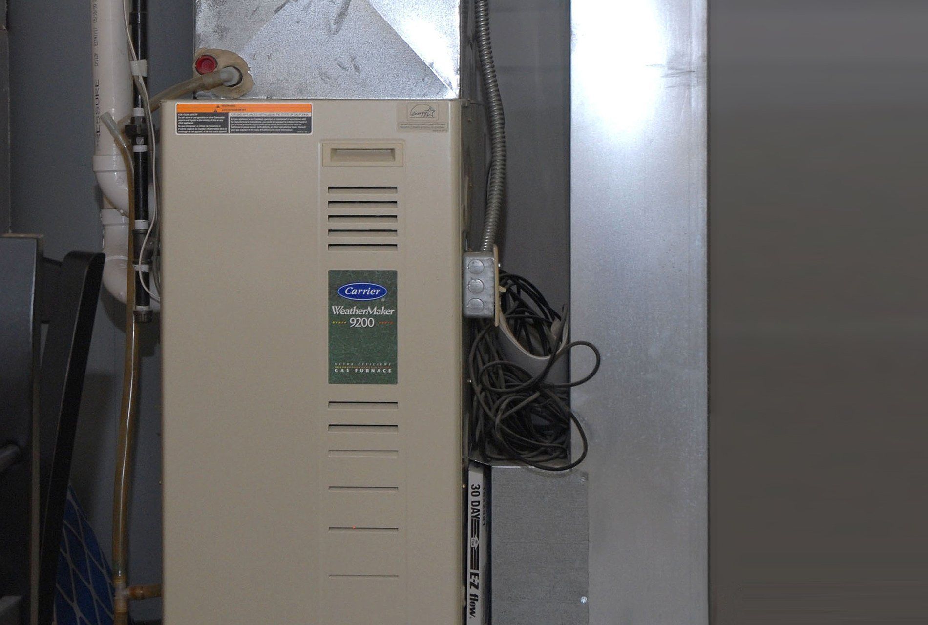 Tan furnace unit in a basement, with silver ductwork and electrical wiring.