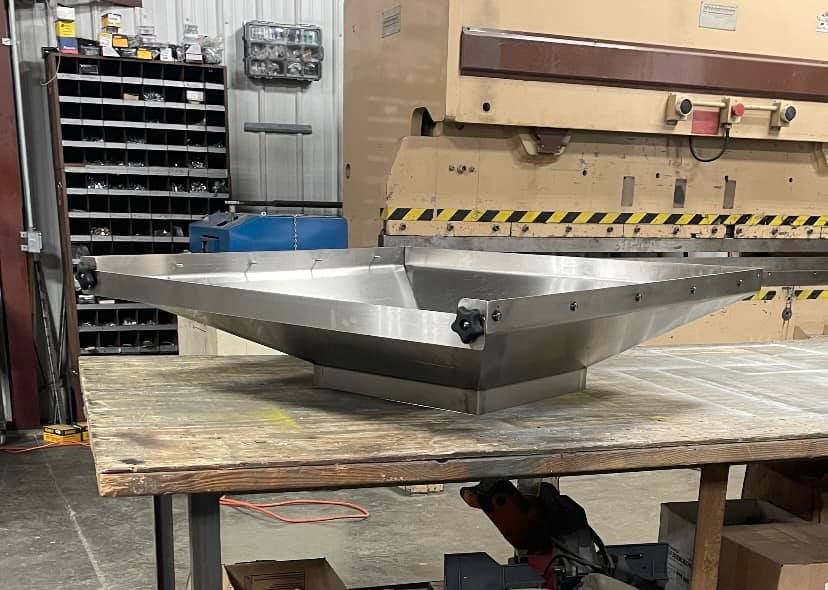 Stainless steel metal hopper on a wooden table in a workshop, with a large press in the background.