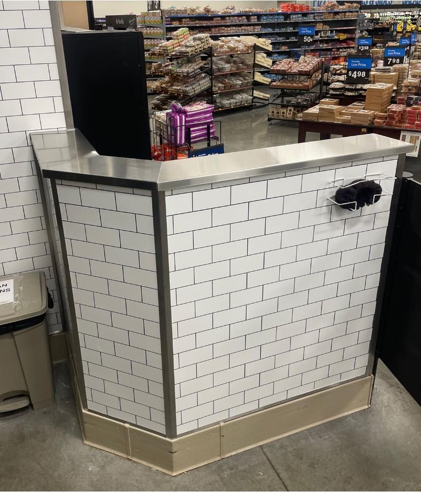 A small counter in a grocery store. White subway tile, metal trim, stainless steel top. Black item on the right side.