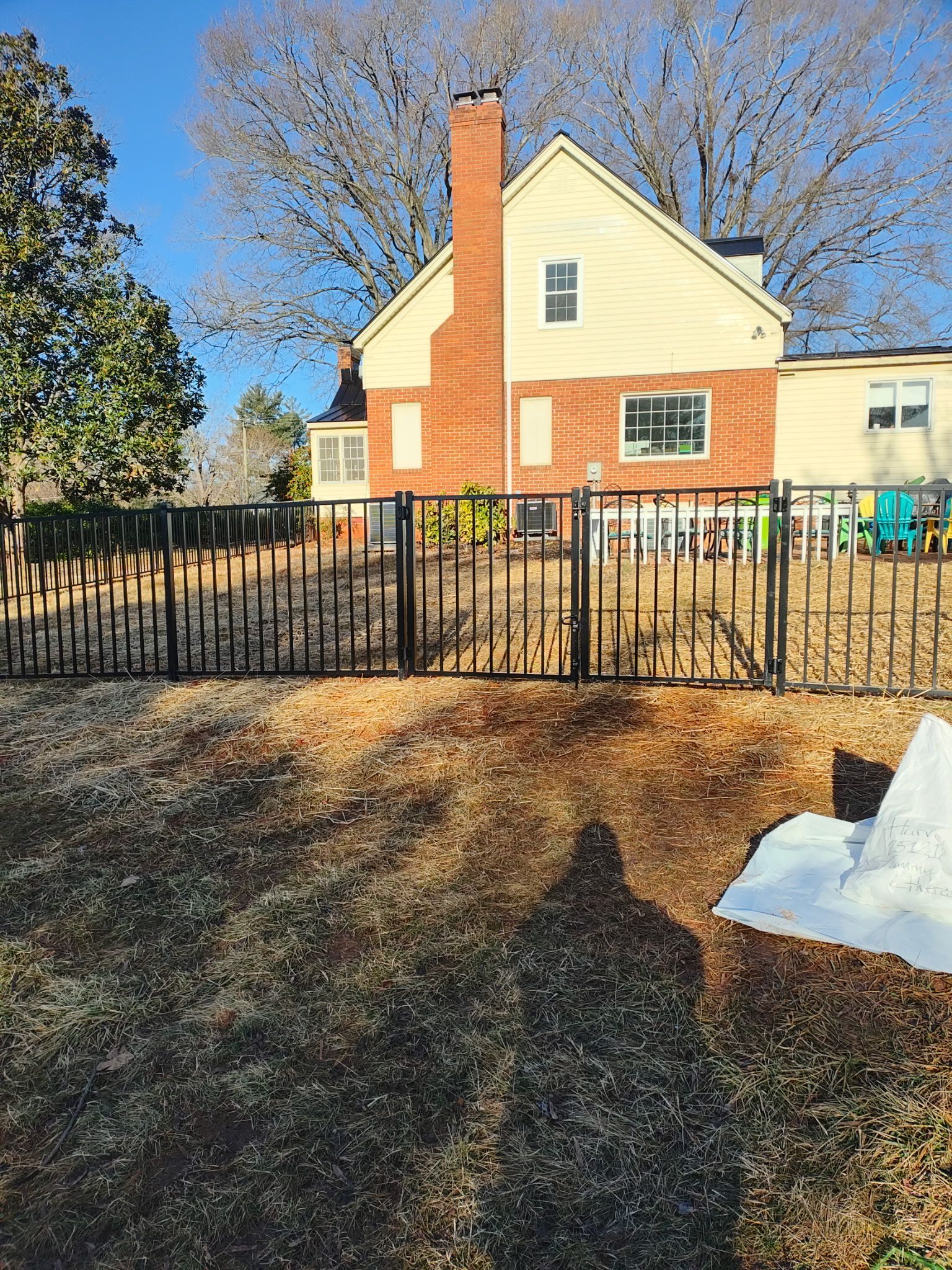 Black metal fence in front of a yellow house with a brick chimney. Dry grass in foreground, blue sky.