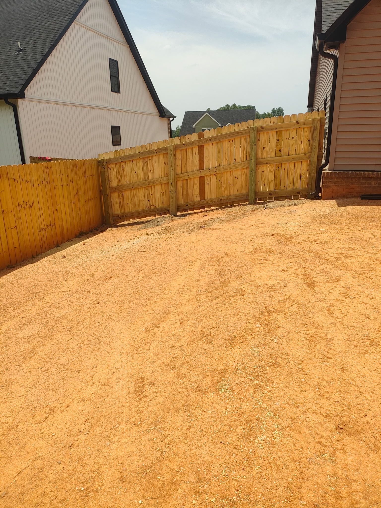 Wooden fence next to a gravel area, with a house visible in the background on a sunny day.