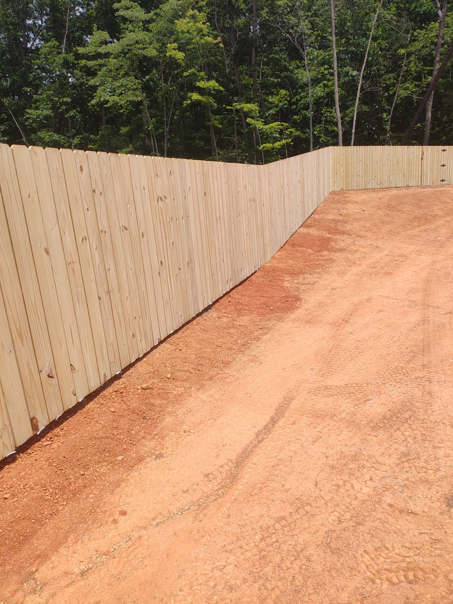 A tan wooden fence borders a red dirt road with green trees in the background.