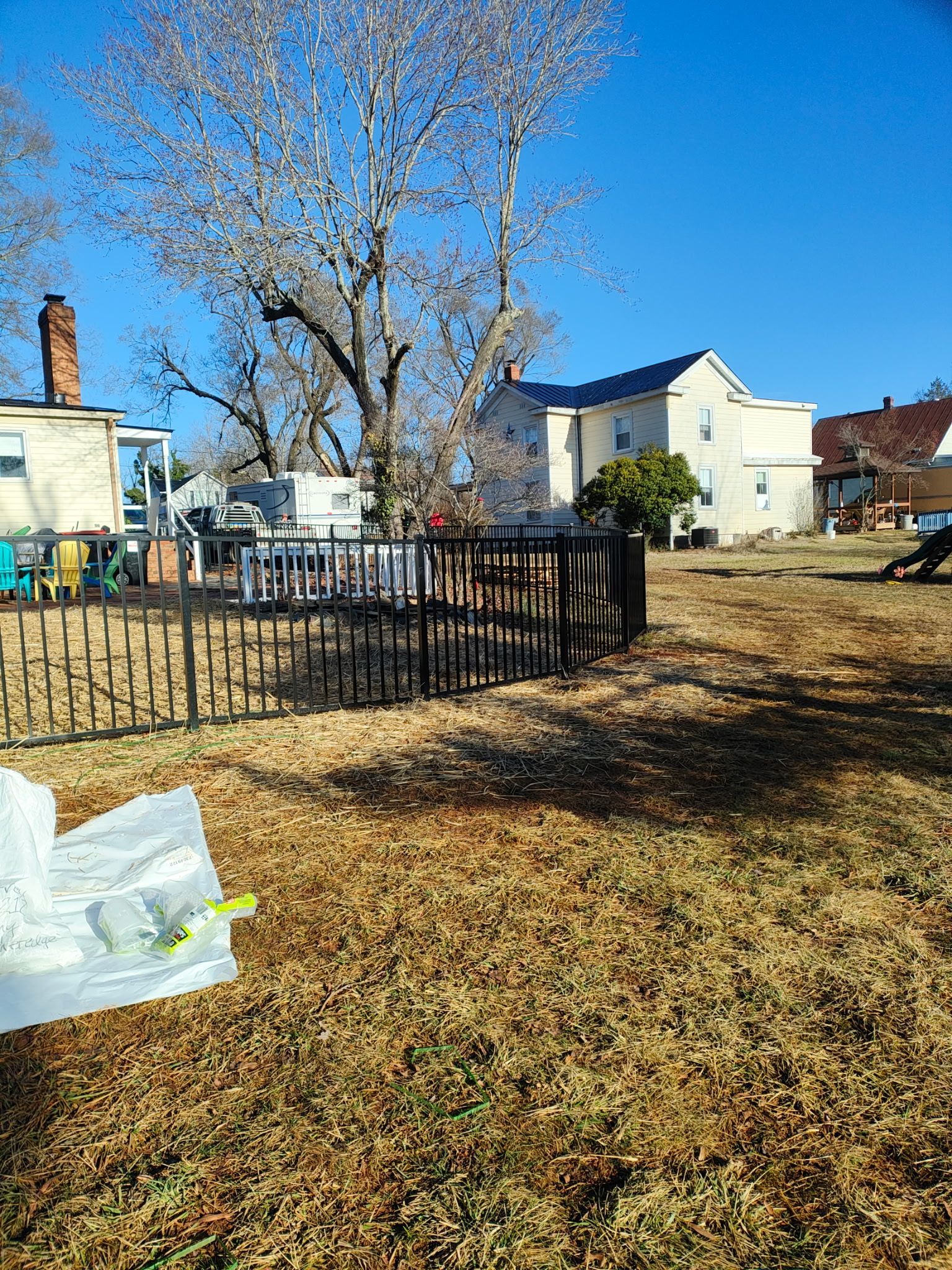 Yard with fence, brown grass, trees without leaves, and buildings under a blue sky.