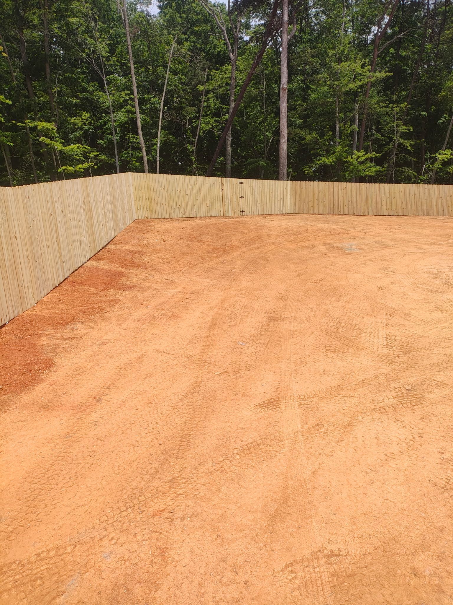 A level red dirt area enclosed by a wooden fence, with trees in the background.