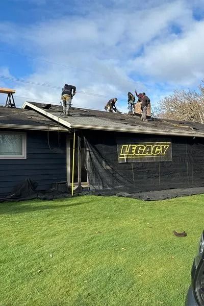 Roofers removing old shingles from a dark-colored house. Green grass and blue sky. 