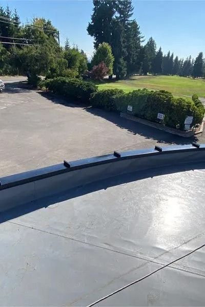 View from a flat roof overlooking a parking lot, bushes, and a golf course under a clear blue sky.