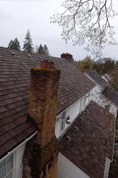 Brown shingled roof with a brick chimney covered in moss, cloudy sky above.