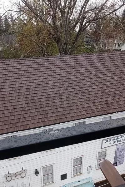 Brown shingled roof over a white building with windows. Trees and overcast sky in the background.