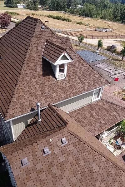 Brown shingle roof of a house with a dormer and vents, set against a green, rural landscape.