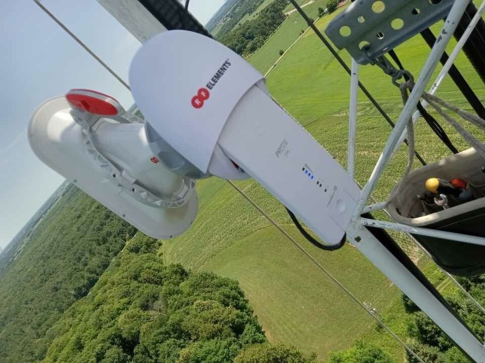 White and gray telecommunications equipment on a tower, overlooking a green field and trees.
