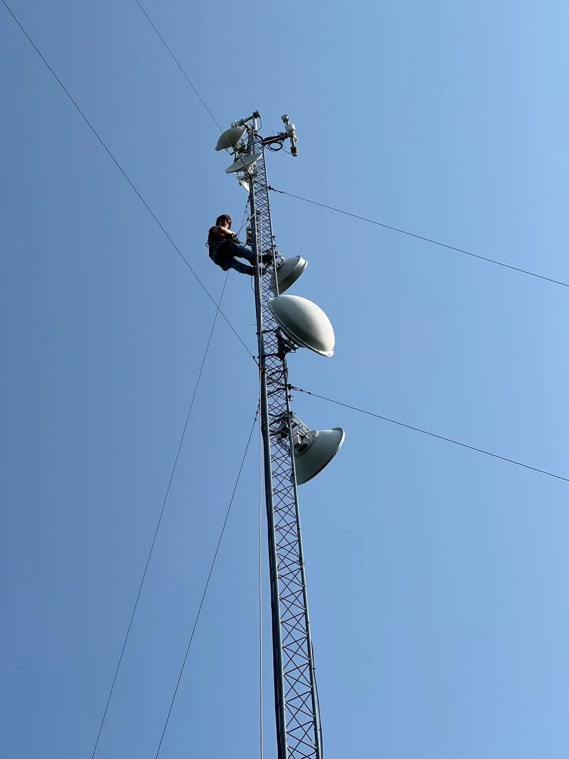 Person climbing a tall communications tower with antennas against a blue sky.