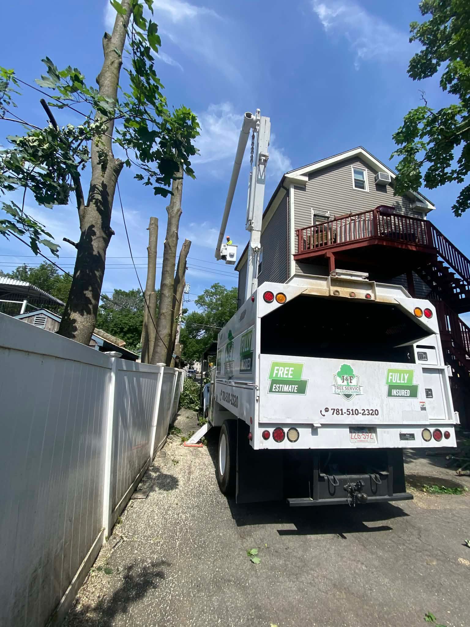 Utility truck in narrow driveway beside fence, carrying a house section on a crane under a blue sky.