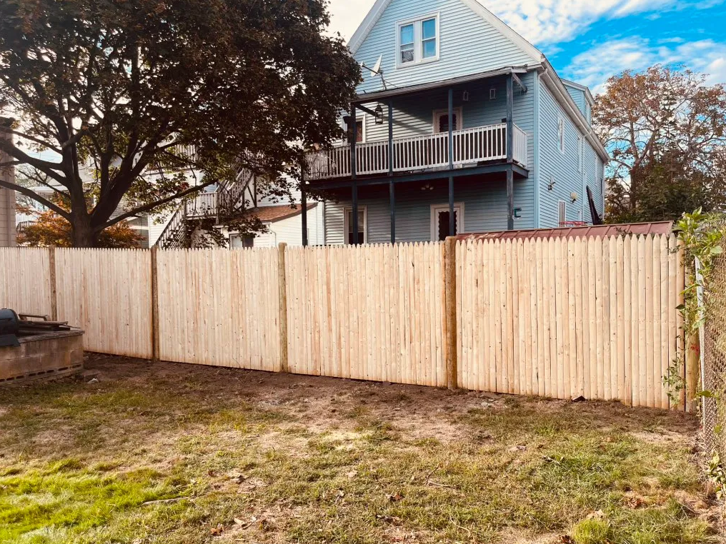 Backyard with wooden fence, patchy grass, and a blue house with porch under trees