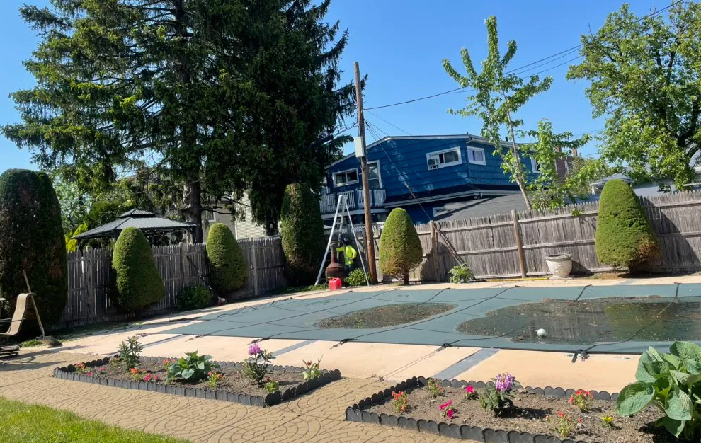 Backyard with a swimming pool, flower beds, tall trees, and a blue house behind a wooden fence