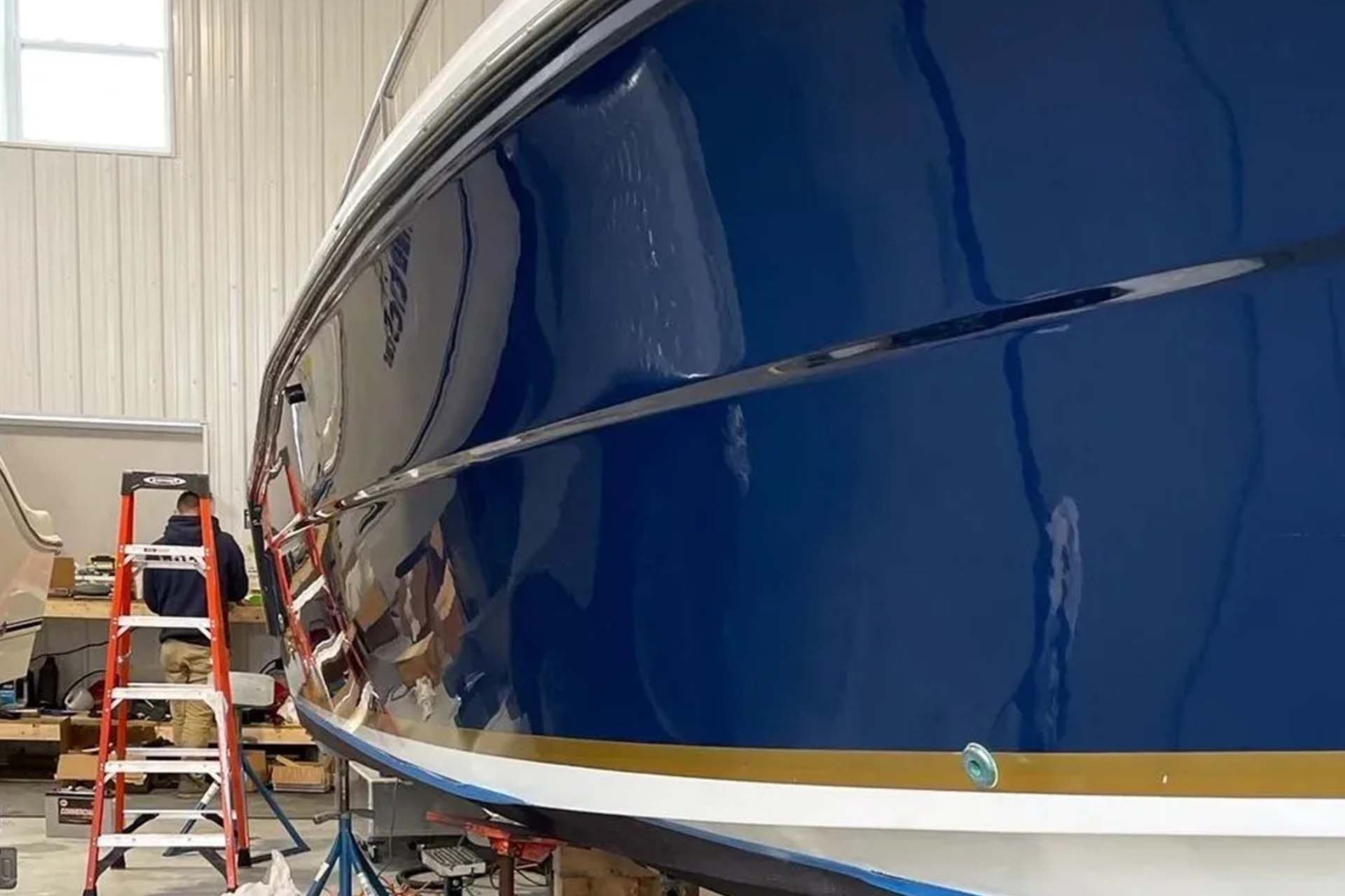 A glossy dark blue boat hull being worked on in a workshop with a ladder visible in the background.