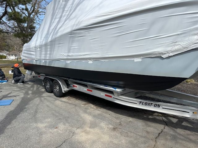 Boat on a trailer, covered in white tarp. Person kneeling, working on the boat's side. Asphalt driveway.