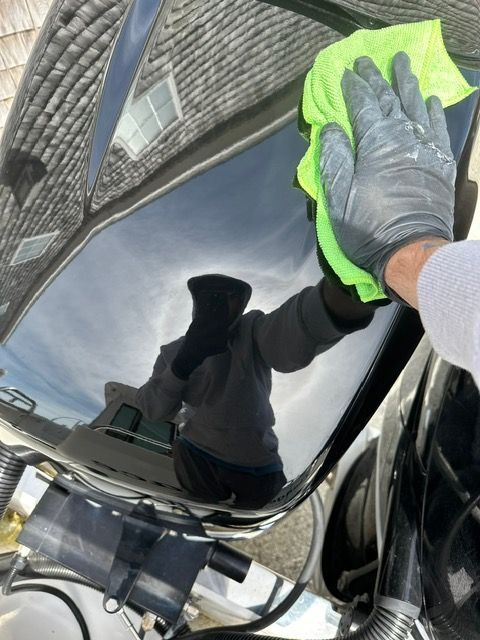 Person in black gloves wiping a shiny black surface with a green cloth; reflection of person and sky visible.