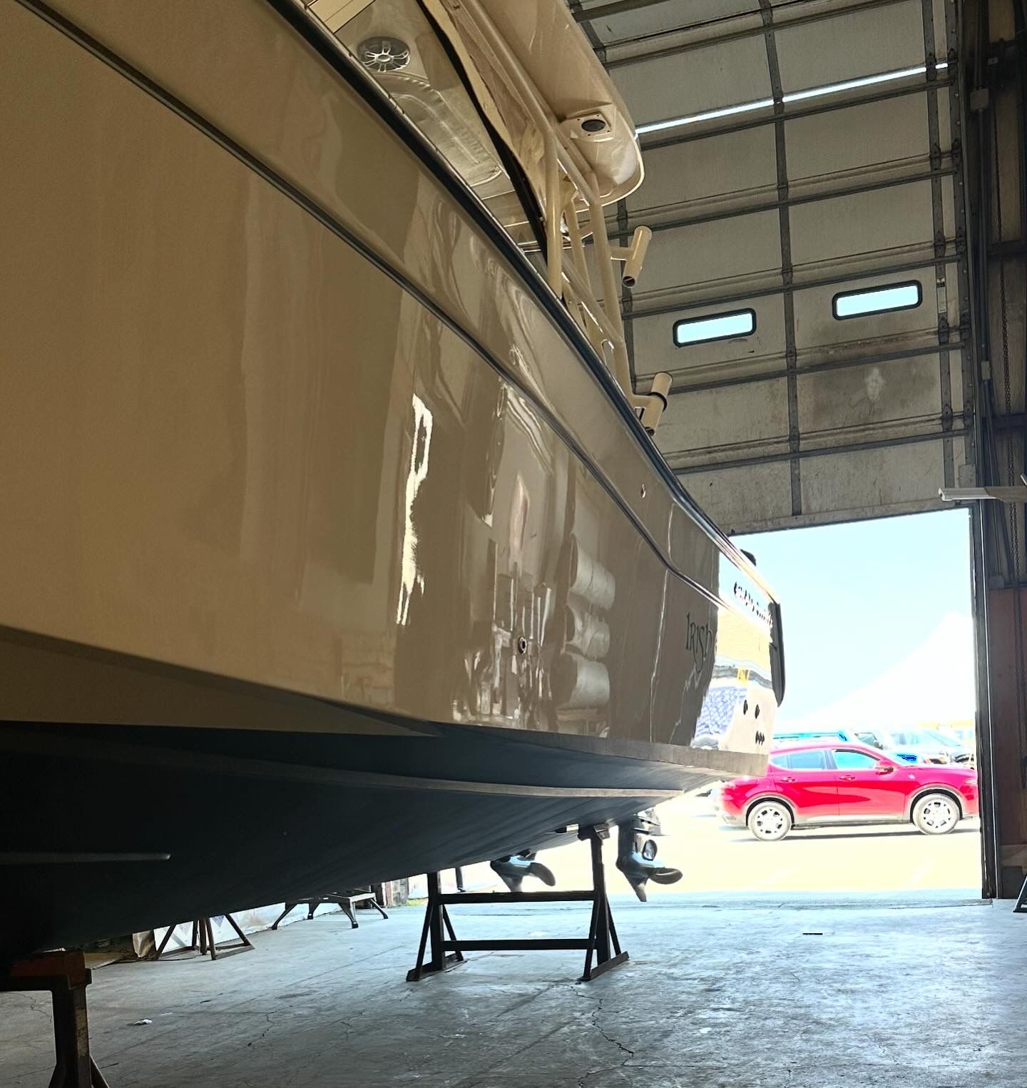 Boat in a maintenance shop, beige hull, blue trim, supported by stands, open garage door, red car visible.
