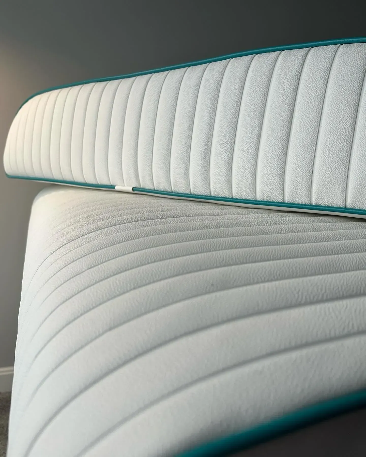 Two white mattresses with teal trim, one stacked on top of the other, against a gray wall.