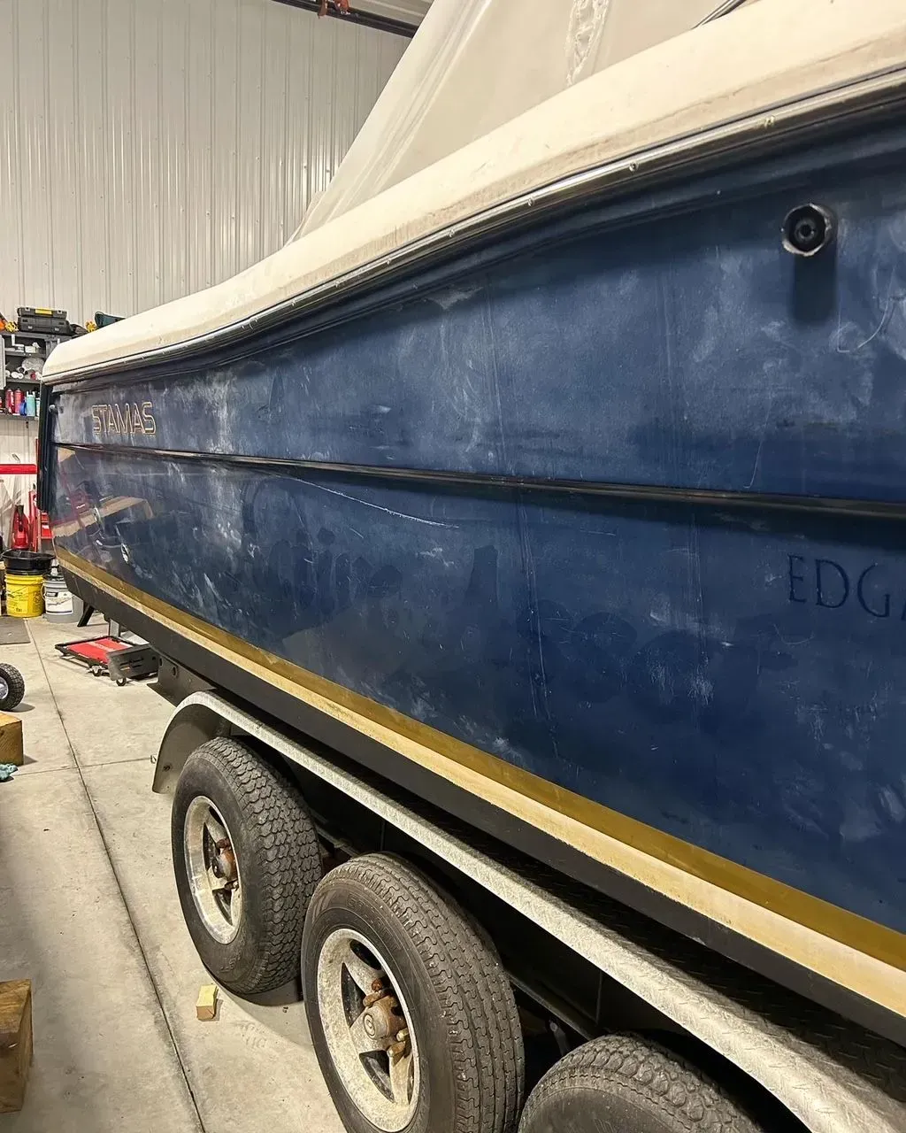 Blue boat on a trailer, in a garage. Boat has a tan trim and white covering. Three tires are visible.