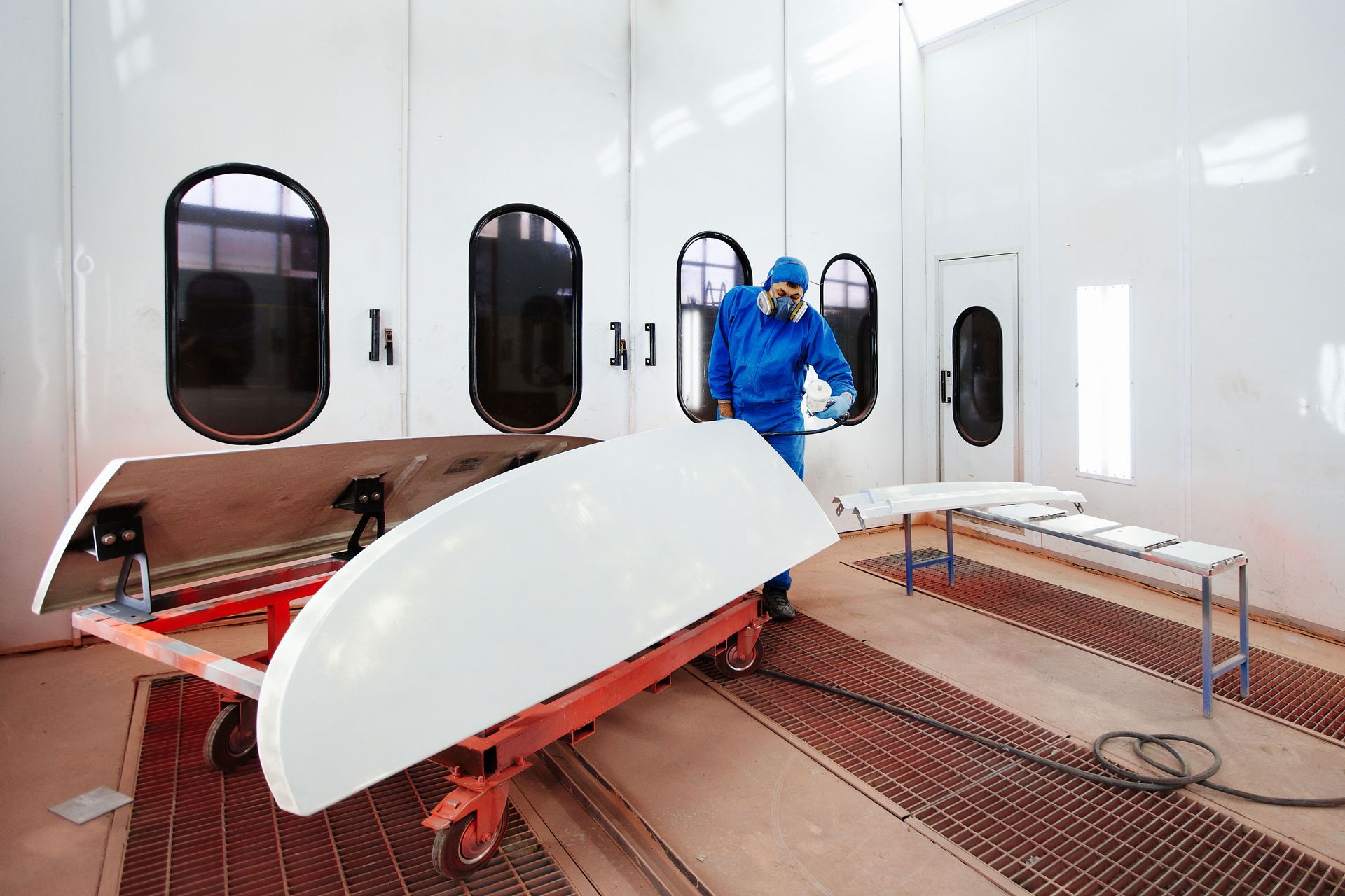 A person in a protective suit painting a large white vehicle part inside a bright spray booth.