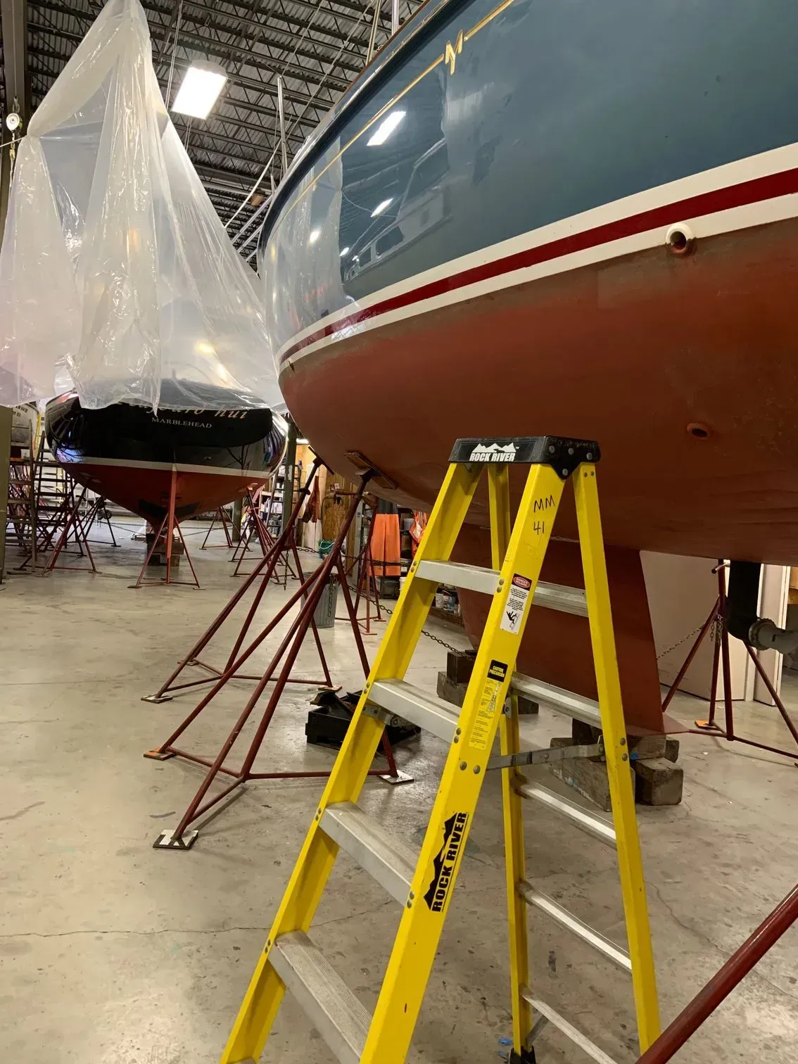 A yellow stepladder stands in a workshop before a blue and red boat hull on a stand, with another boat covered in plastic.