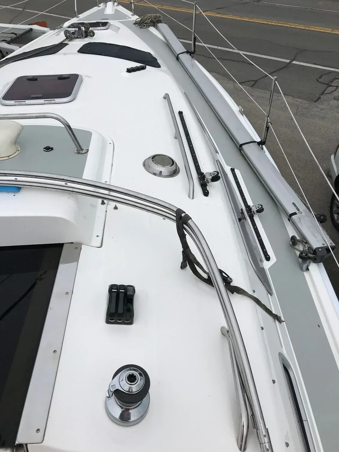 The deck of a white sailboat showing a winch, rope clutch, handrail, and track hardware along the starboard side.