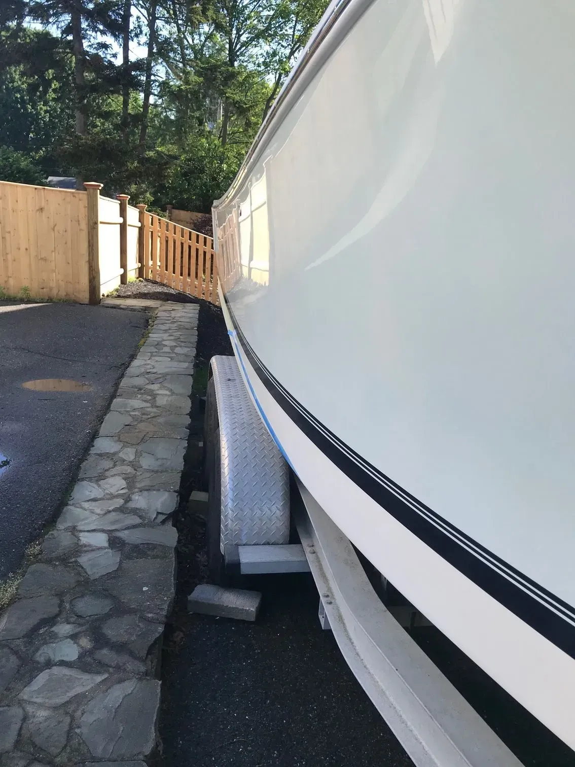 A white boat on a trailer parked beside a stone walkway and wooden fence.