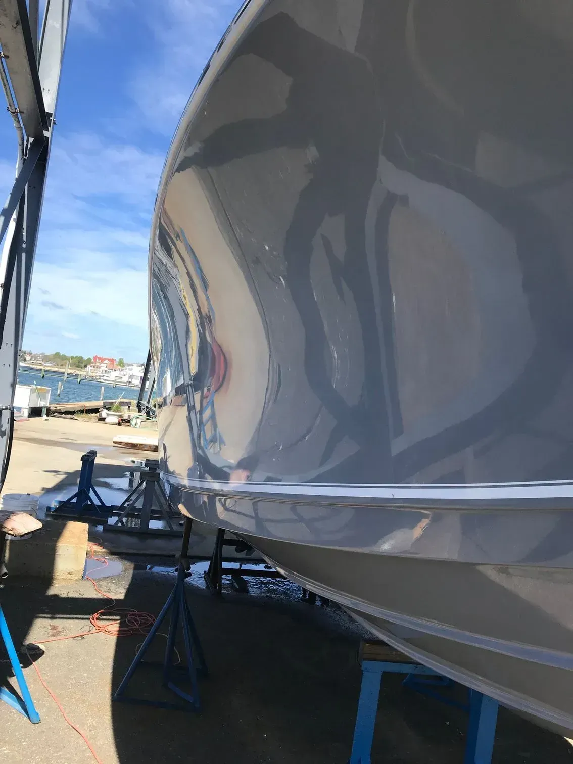 A glossy, reflective gray boat hull rests on metal stands in a shipyard beside a harbor under a sunny blue sky.