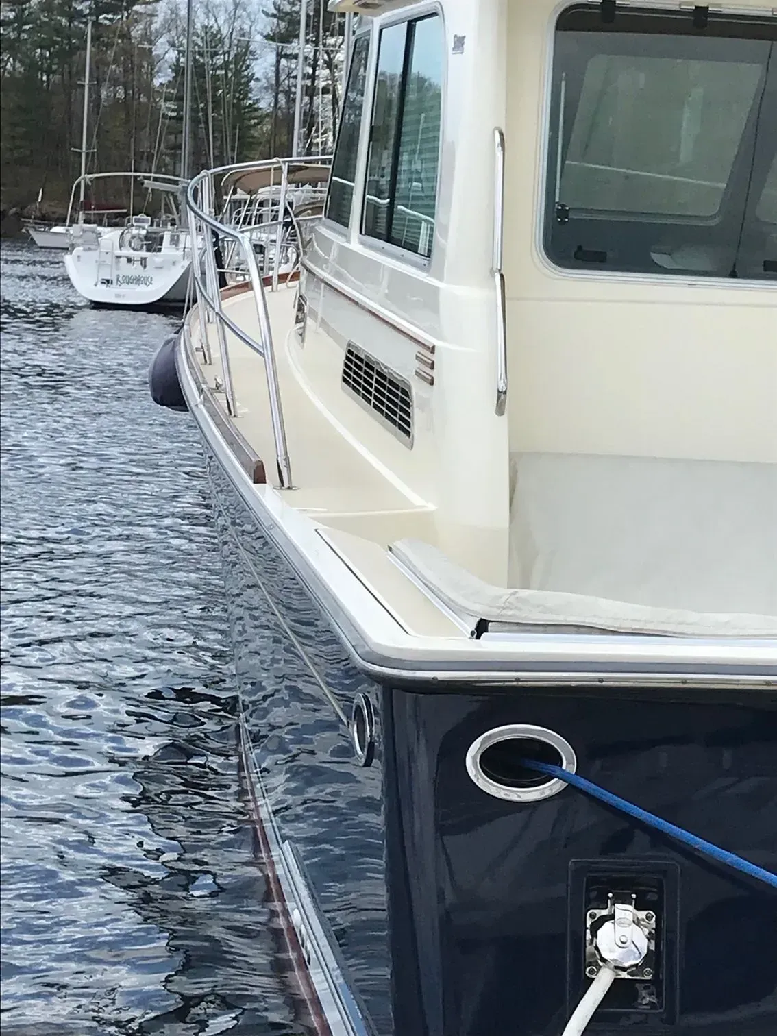 The stern and starboard side of a boat with a dark blue hull and cream cabin, moored near another boat in calm water.