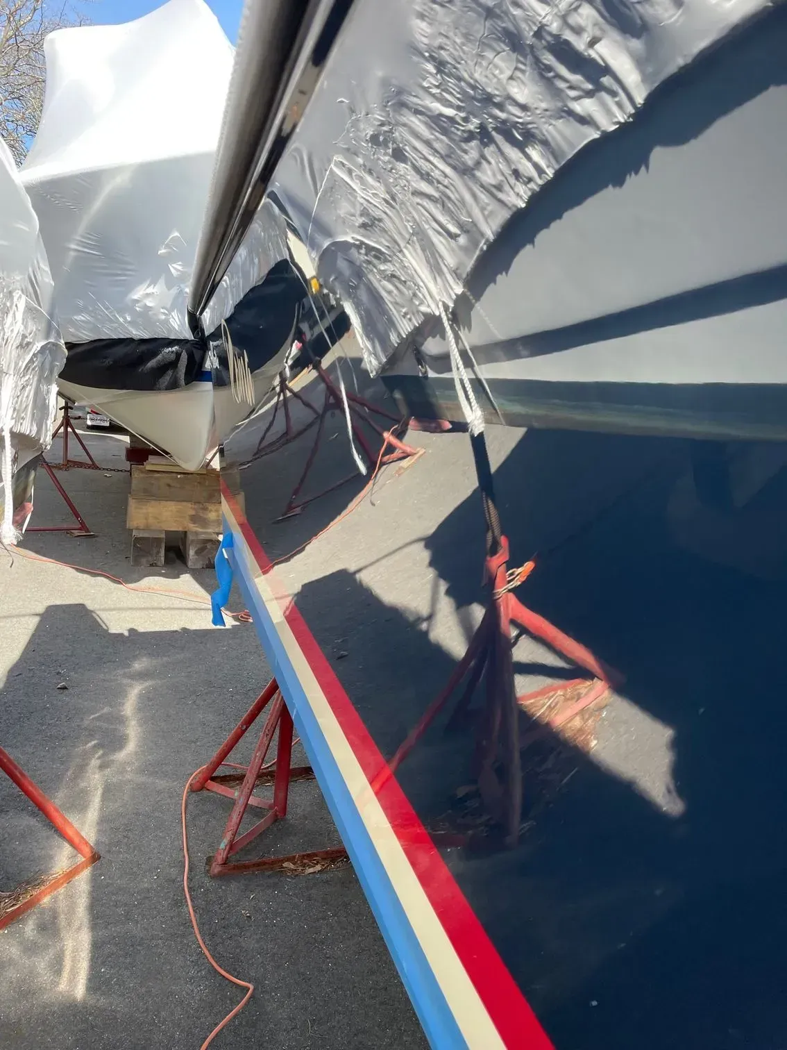 A low-angle view of a boat hull in dry dock, supported by red metal stands, with a tarp partially covering the deck.