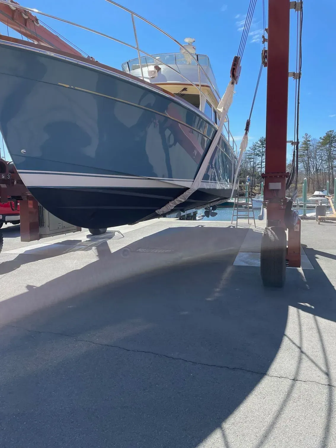 A boat with a dark hull and light upper structure being lifted by a large red travel lift in a boatyard.