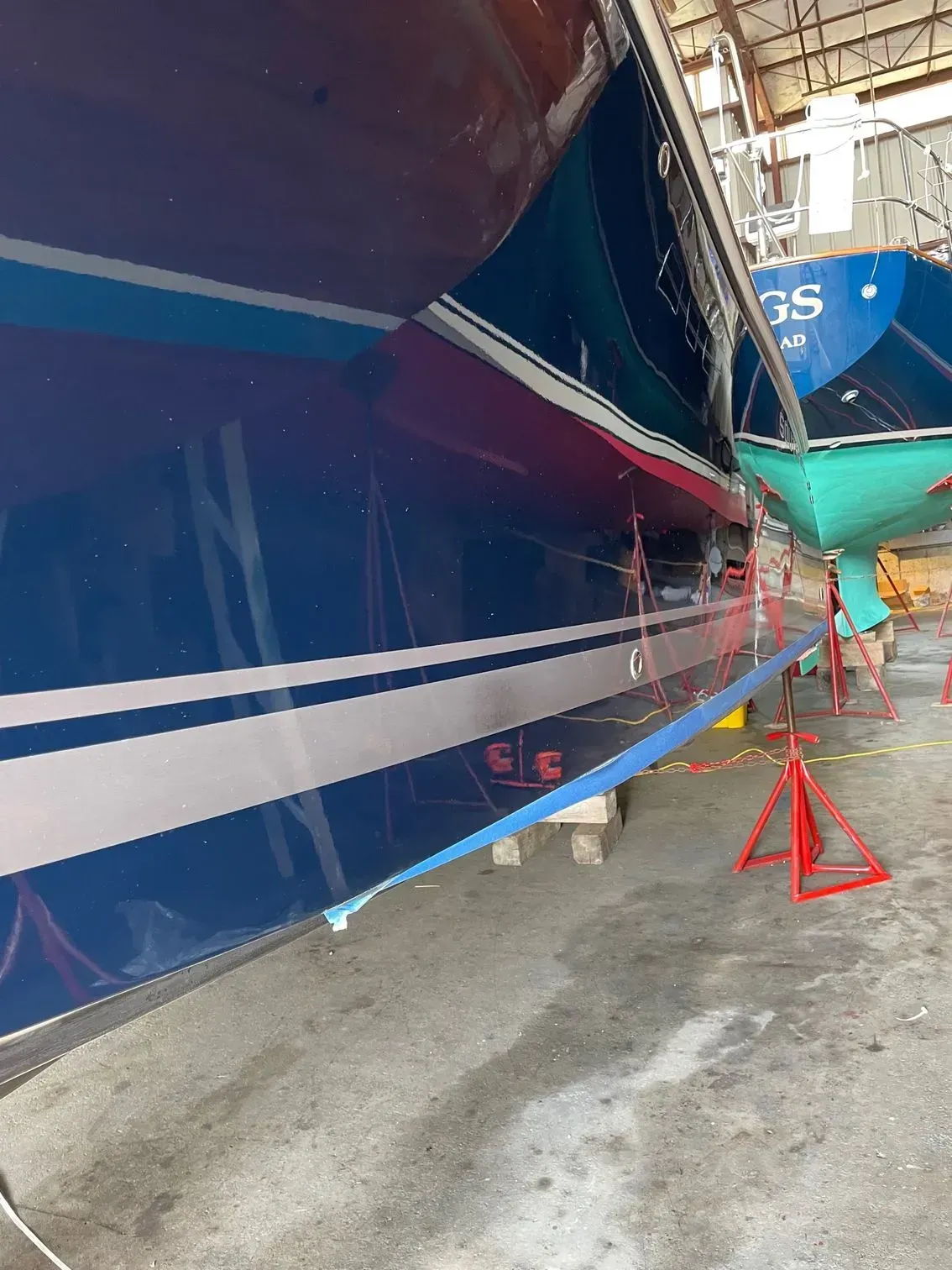 A dark blue sailboat hull in dry dock, showing a light blue stripe and a red metal support stand on a concrete floor.