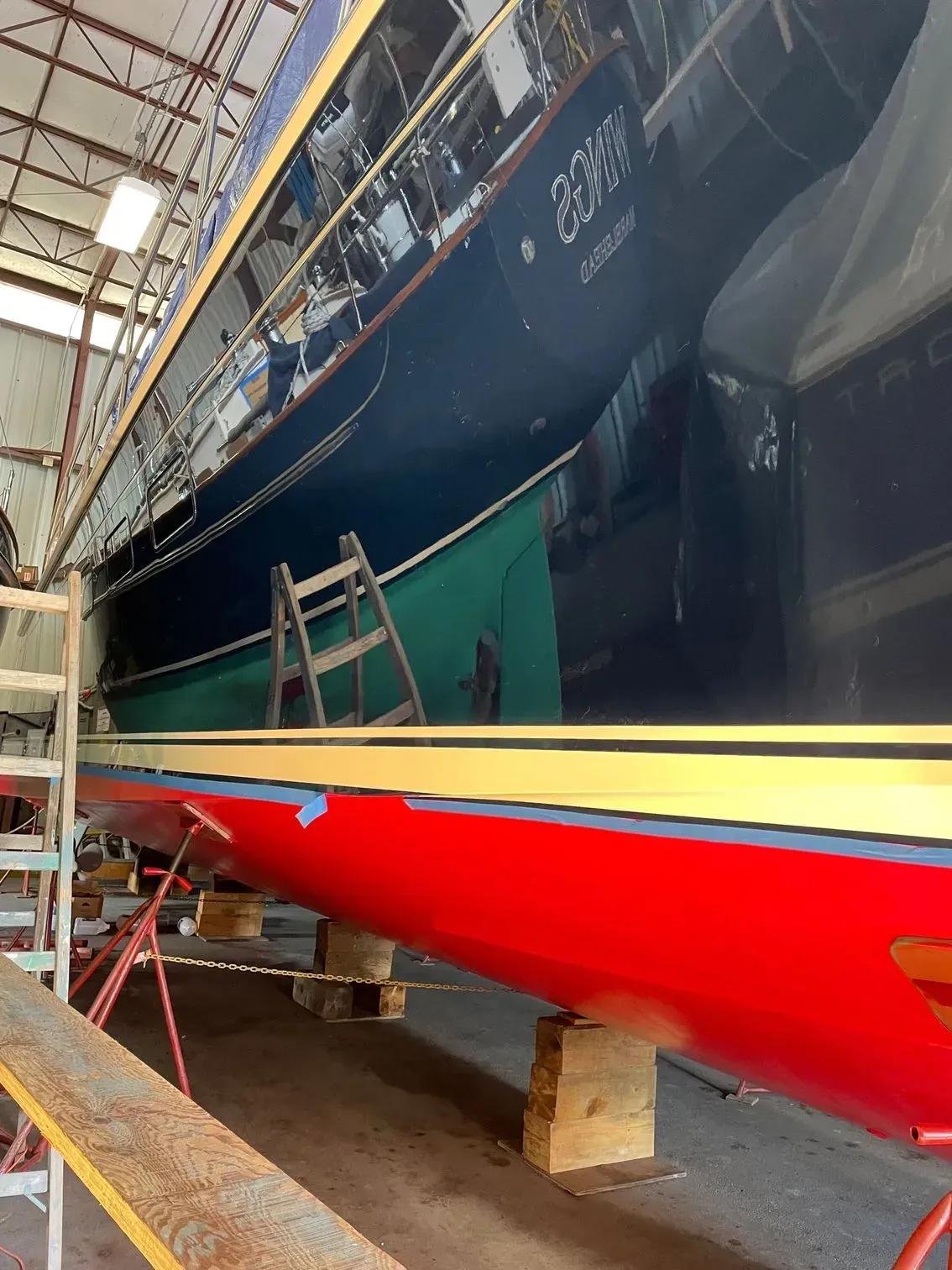 A large dark blue boat sits in a workshop above another boat with a bright red hull, both supported by wooden blocks.