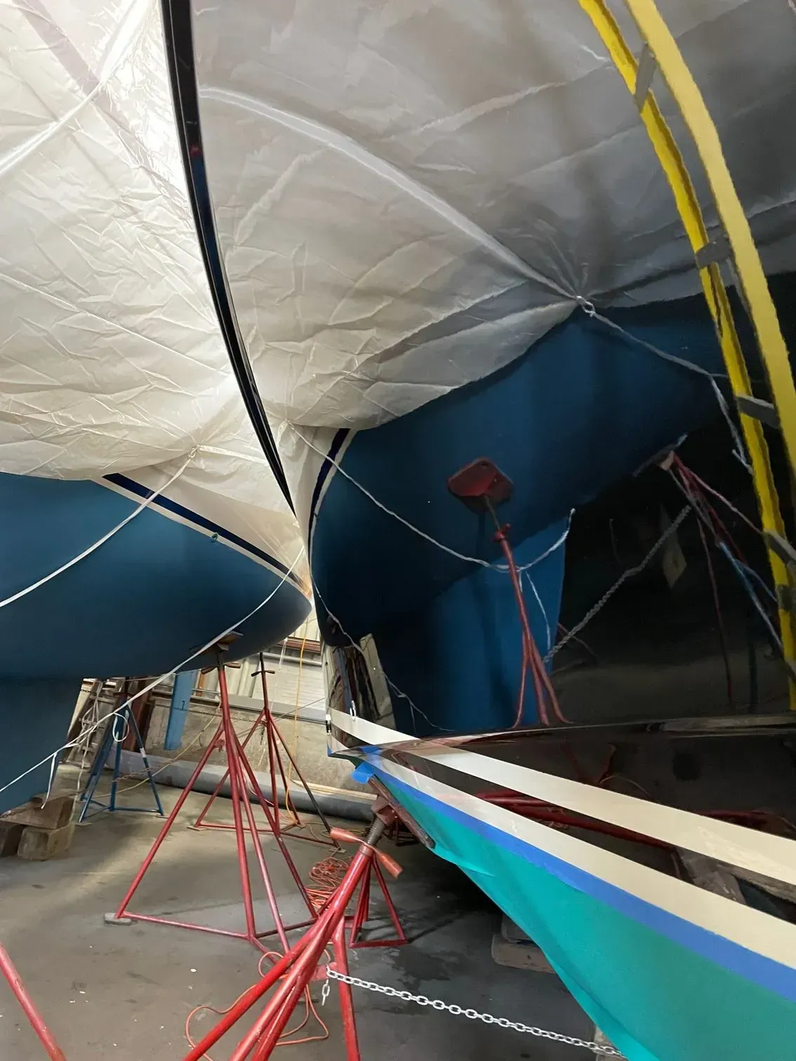 Two blue sailboat hulls under protective covers in a workshop, supported by red metal stands and ladders.