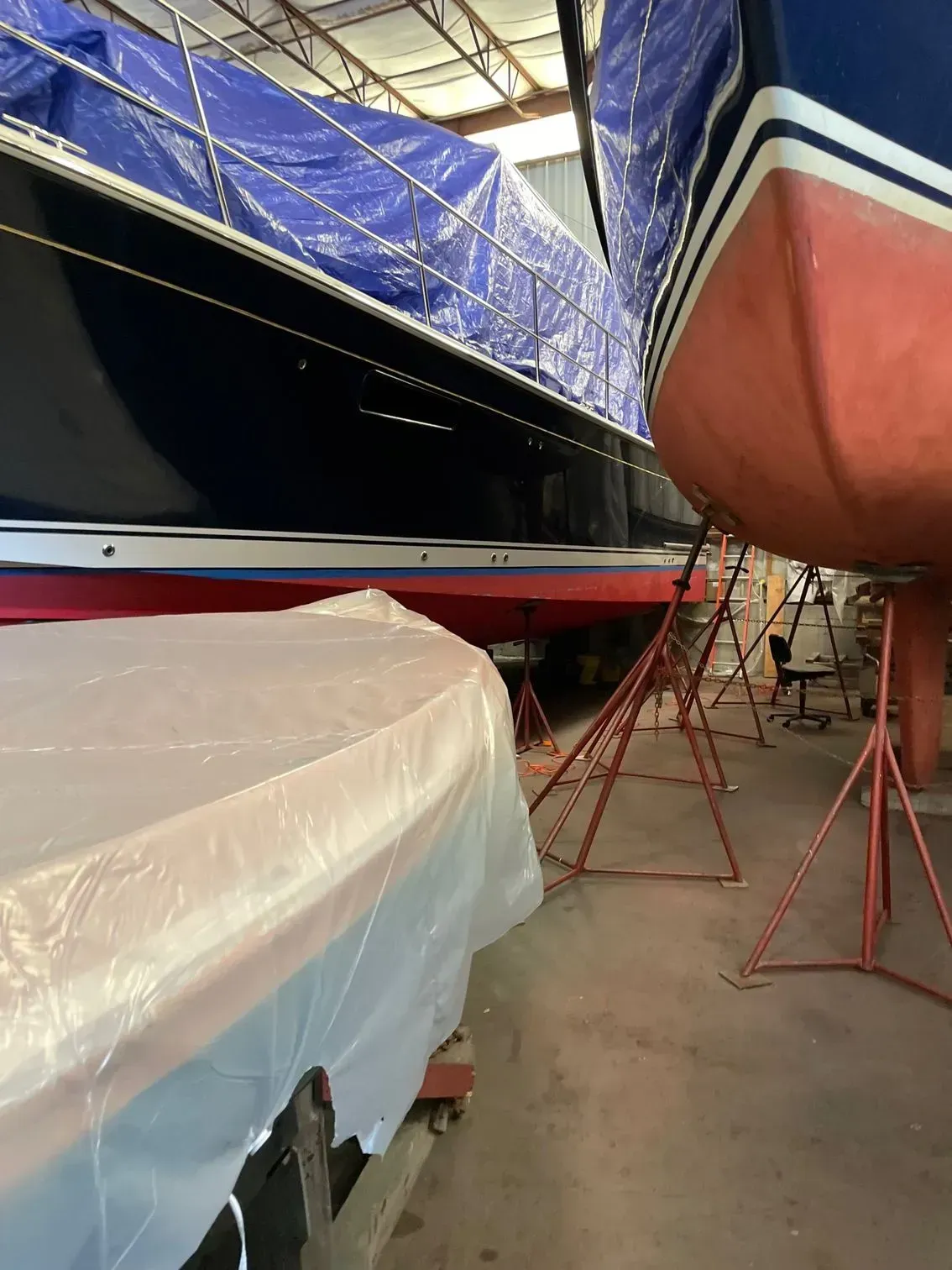 Boats stored in an indoor facility, with one covered in blue tarp, one in plastic, and one showing a red hull.